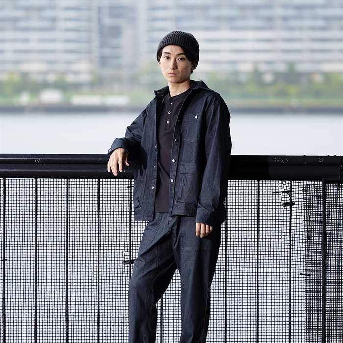 ヘンリーネックTシャツにストレッチデニムのジャケットとパンツを合わせ、手すりにもたれる男性